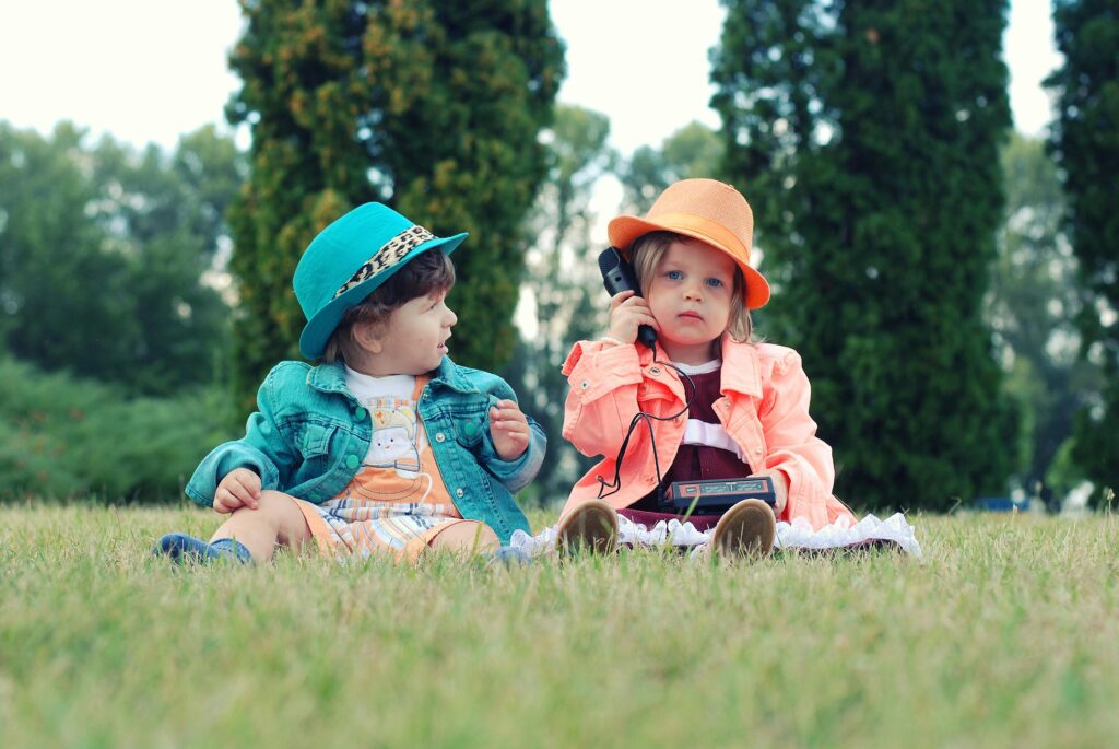 Two toddlers in colorful outfits enjoying playtime with walkie talkies in a sunny park setting.